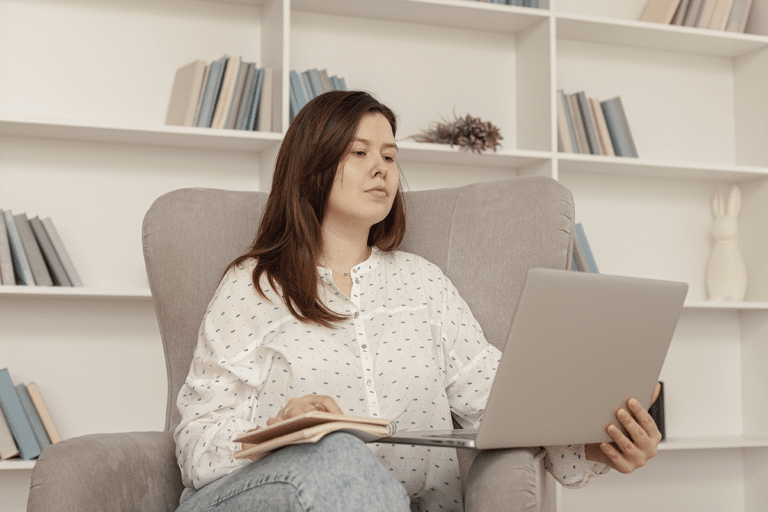 Woman sitting on a sofa with a book while looking thoughtfully at her laptop, illustrating the feeling of guilt when trying to rest