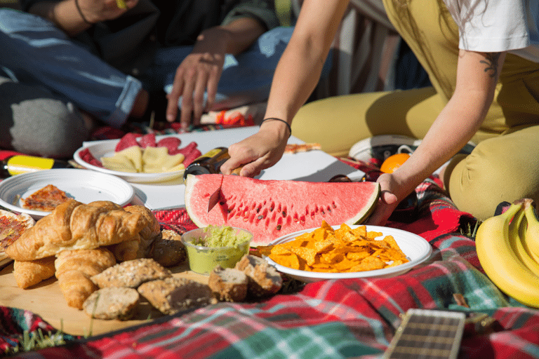 Friends sharing watermelon, bread, and snacks at a sunny outdoor picnic