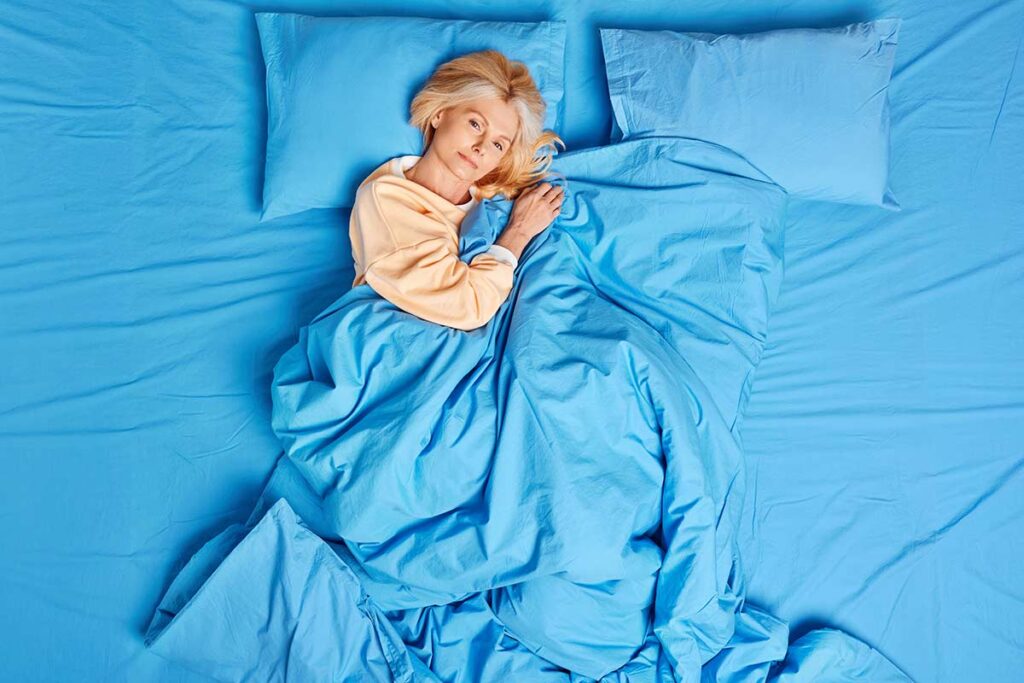 Woman in her 50s resting under blue bedding, illustrating cooling blankets for menopause night sweats and hot sleepers